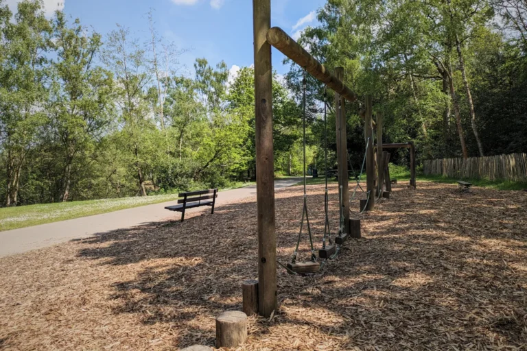 Outdoor park with wooden balance playground equipment, benches, and a paved path surrounded by trees