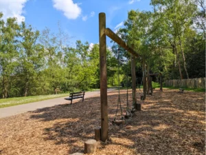 Outdoor park with wooden balance playground equipment, benches, and a paved path surrounded by trees
