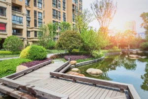 Landscaped residential garden with a wooden bridge over a pond, surrounded by greenery and apartment buildings in sunlight